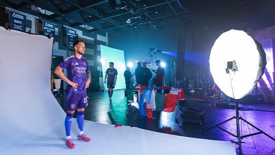 An Orlando City player poses for the camera with their hands on their hips wearing a purple uniform. Production crew members stand around in the background.