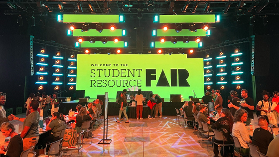 Students and staff sit at tables and interact. A large screen behind them displays “Welcome to the Student Resource Fair” in black text with a green background.