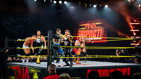 Wrestlers duke it out in the ring during a TNA Impact wrestling match, surrounded by bright stage lights and a cheering crowd in Full Sail's Live Venue.