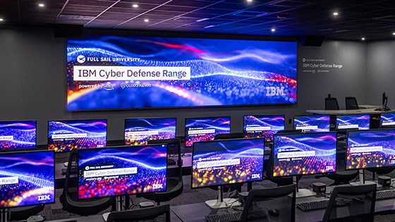 A room with dozens of desktop computers and chairs at long tables. A large screen on the wall reads Full Sail University IBM Cyber Defense Range Powered by AWS and Cloud Range.