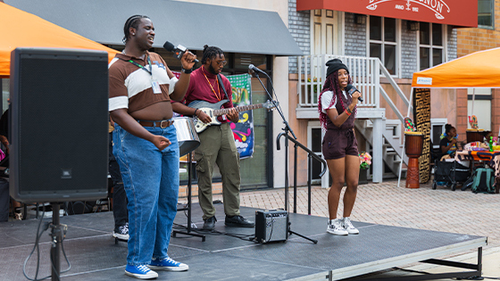 Three performers stand onstage. Two are holding microphones and singing, and one is playing an electric guitar