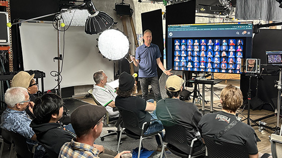 Shawn Rinehart stands in front of an audience of students and faculty next to a screen displaying numerous headshots. Lighting and camera equipment are around the room.