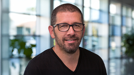 Sebastian Krys smiles at the camera in a lobby with large glass windows. He is wearing glasses and a black t-shirt.