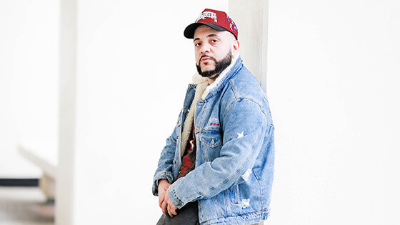 A man wearing a red baseball cap and a denim jacket is pictured leaning against a concrete pillar in a brightly lit room.