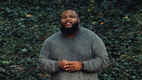 Tariq Ricks poses for the camera in front of a green leaf background. He is wearing a long sleeve grey shirt.