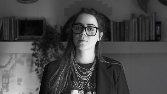 Veronica Concha stands in front of a bookshelf in her home. She is wearing a dark blazer, a band t-shirt, silver necklaces, and glasses.