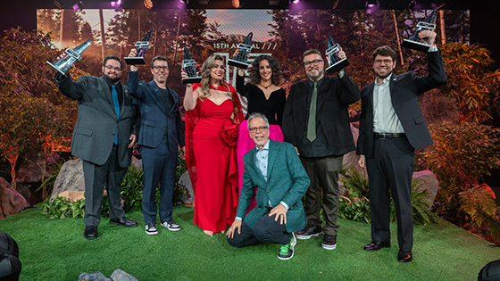 The Hall of Fame 15 inductees raise their awards in the air on a stage decorated with foliage. Garry Jones kneels in front.