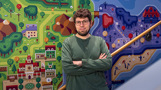 A bearded man in glasses and a green shirt stands with his arms crossed in front of a colorful, game-inspired illustrated mural.