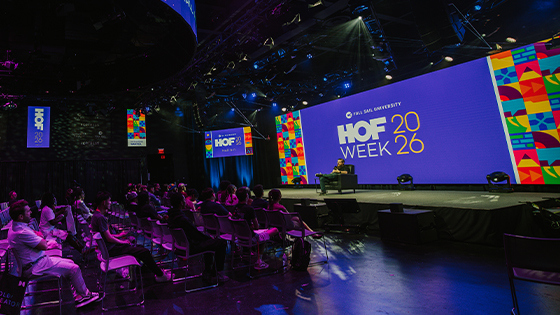 A speakers sits in a chair on a stage with a large screen behind them displaying “HOF Week 2026” and colorful graphics. An audience sits in rows of chairs facing the stage.