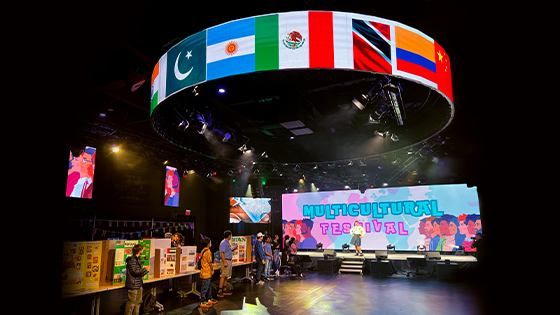 Students stand by a line of tables against a wall with cardboard presentations on various countries. One student poses onstage in a kilt and white shirt with a large screen behind them displaying “Multicultural Festival.” Above the room, a circular screen displays flags of various countries.