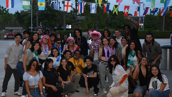A diverse group of young adults poses outdoors under international flags, dressed festively with leis and fun accessories.