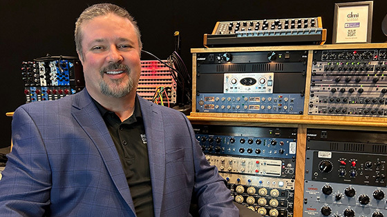 Richard Tremmel smiles at the camera with an array of music recording equipment behind him. He is wearing a black shirt and blue blazer.