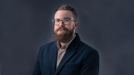 Drew Bailey wears a blue blazer and a brown checked shirt. He has brown hair, a beard, and glasses, and he’s smiling at the camera.