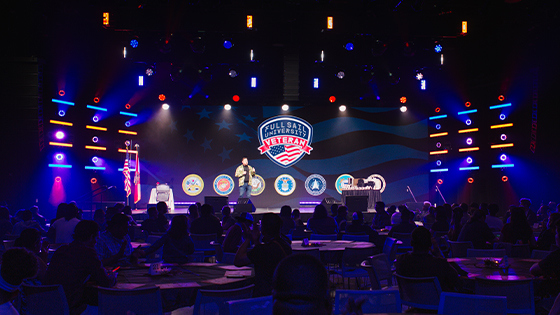 A crowd of people sit at round tables in a large room. A speaker stands on a stage holding a microphone with a screen behind them displaying the Full Sail University Veteran logo and logos of the branches of the military.