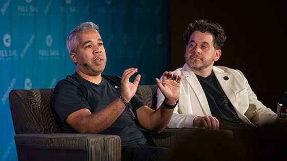 Juan Peralta and Gary Rizzo sit in armchairs on a stage and give a presentation. Juan wears a black t-shirt and Gary wears a black shirt and white blazer.