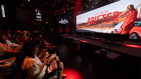 Students pack the rows of a live venue, watching a large screen presentation for ‘Drifter,’ with red stage lighting and a panel setup on stage.