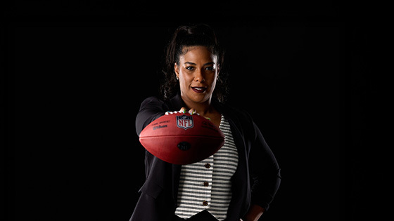 A woman in a blazer holds an NFL football toward the camera, confidently posing against a dark backdrop.