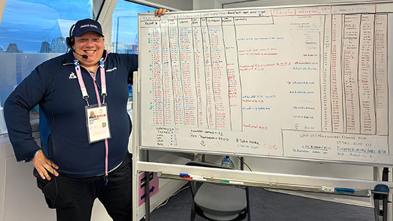 Alberto Pisani stands next to a large whiteboard with flight numbers on it and smiles. He wears a headset and a baseball cap that says Paris 2024.