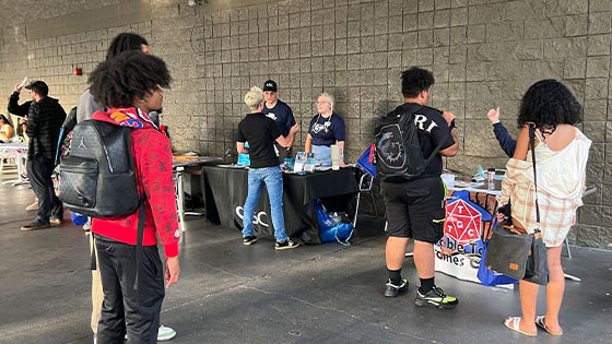 Several students gather on a patio with various tables set up. Club representatives stand at the tables talking with students.