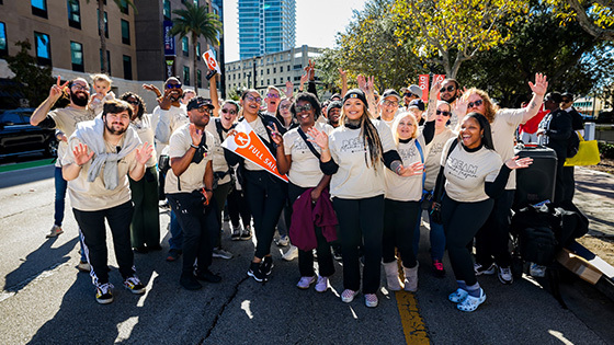 A large group of staff and students smile and wave at the camera wearing matching beige t-shirts. Buildings and trees are behind them.