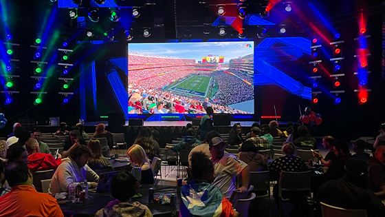 A crowd of students sit at round tables in a large room facing a large screen displaying a football stadium. One student wears a Puerto Rican flag as a cape.