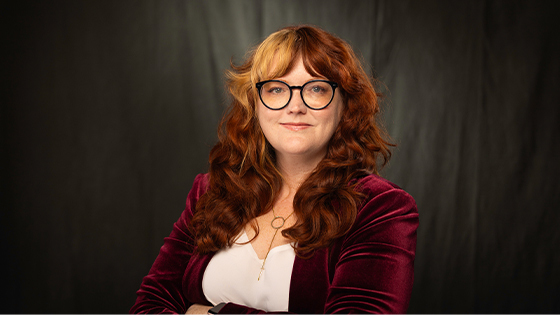 Susan Kost smiles at the camera. She has long red hair and is wearing a white blouse, a red velvet jacket, and black glasses.