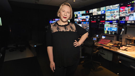 A person with blonde hair and a black lace-accent top stands in a dim control room, smiling, with colorful broadcast screens glowing behind them.
