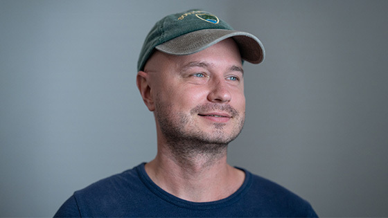 A portrait of a man wearing a green baseball cap and navy shirt while looking off to the side against a grey-green backdrop.