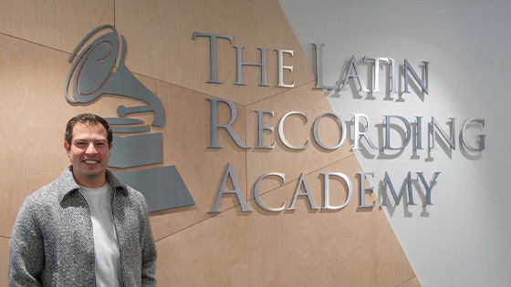 Nahun Paguada stands in front of a wall with a large sign displaying “The Latin Recording Academy” with a gramophone logo. He is wearing a white shirt, grey jacket, and khaki pants with his hands in his pockets.