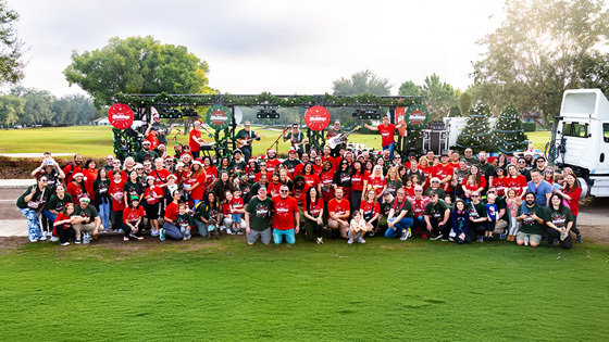 A large group of staff, faculty, and their families pose for a photo wearing red and green holiday shirts in front of a semi-truck bed decorated with garlands and lights. Some stand on the truck bed holding various musical instruments.