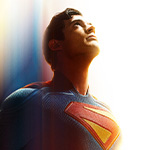 Superman stands and looks at the sky. He is wearing his blue costume with a yellow and red S on the front and his red cape is billowing behind him.