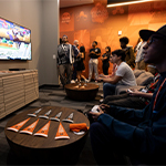 Gamers compete in a lounge with dual screens, a PlayStation 5, and Armada-branded decor, while a crowd watches in the background.