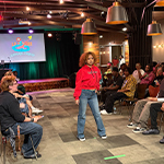 A student walks the runway wearing a red hoodie and jeans. The audience sits in two rows of chairs on either side of the runway facing the model. A DJ stands on the stage behind the runway.