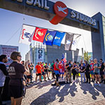 Three 5K winners stand on a podium with medals around their necks under the Full Sail Studios archway. Other competitors stand near them and applaud.