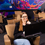 Students study and relax with laptops in Full Sail’s 3B Art Building Lobby, featuring colorful mural art and lounge seating.