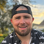 Tyler Yates sits on a bench in an outdoor park and smiles. He’s wearing a black and white collared shirt with dinosaurs on it and a backwards cap.