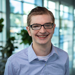 Wes Locher stands in a lobby with lots of windows and smiles at the camera. He wears glasses and a blue checked button-up shirt.