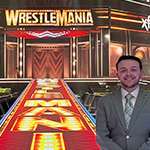 Colby Applegate wears a suit and stands in a brightly lit wrestling arena with Wrestlemania graphics displayed on the walls and floor.
