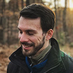 Mikey Zoppo stands in a forest with sunlight poking through the trees behind him. He looks down and smiles, and he is wearing a dark jacket.