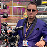 Andrew Karr wears sunglasses and a purple suit as he holds a camera in front of a wrestling ring in a WrestleMania arena.