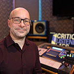 Aaron Gandia sits in a recording studio. He is smiling and wearing glasses and a burgundy shirt. A soundboard is behind him.