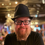 David Dennison sits at a table with his arms crossed. He has glasses and a beard and is wearing a black t-shirt and black fedora.