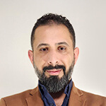 Dr. Ahmed Al Zaidy stands with his arms crossed and smiles at the camera. He is wearing a brown blazer over a blue button-up shirt.