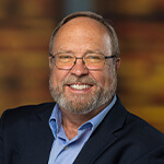 Reo McBride stands with his arms crossed and smiles at the camera. He has a beard and glasses; he’s wearing a blue button-up shirt and a blue blazer.