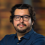 A man with glasses and a beard poses confidently with his arms folded against a warm, blurred background.