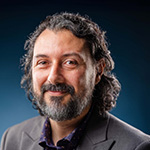 Plinio Pinto smiles at the camera. He has curly brown hair and a beard streaked with gray, and he wears a gray blazer.