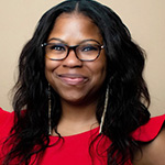 A woman with long, wavy black hair, glasses, gold earrings, and a red dress with ruffled sleeves smiles warmly.
