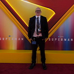 Grad Guillermo Cummings stands before a glowing Superman logo backdrop at a movie premiere, wearing a lanyard and event badge.