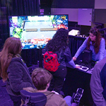 Several people stand around facing a table with adaptive gaming consoles, speakers, and a large screen nearby displaying a game. A staff member stands behind the table.