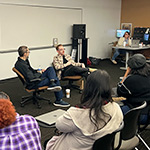 Brad Biwer sits in a chair and speaks to a crowd of students. A staff member sits in a chair next to him and a faculty member sits at their desk behind them.